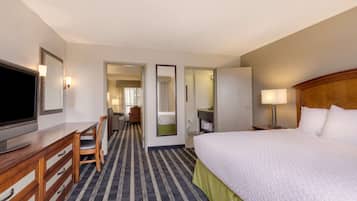 A hotel room with a large bed, a desk with a flat-screen TV, a chair, a mirror, and a bathroom visible through an open door.