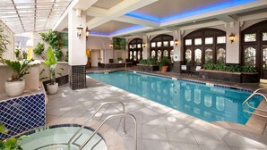 An indoor swimming pool with a tiled floor, a glass-enclosed area with plants, and a poolside seating area.