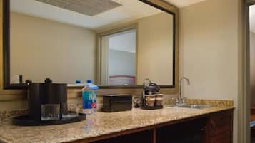 Baño de hotel con encimera de granito, lavamanos, espejo, cafetera, una botella de agua Fiji y un microondas.