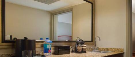 A hotel bathroom with a granite countertop, a sink, a mirror, a coffee maker, a bottle of Fiji water, and a microwave.