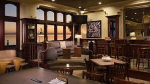 A spacious lounge area with a large window offering a view of the desert landscape, a comfortable seating area with a sofa and armchairs, a dining table with chairs, and a ceiling fan.