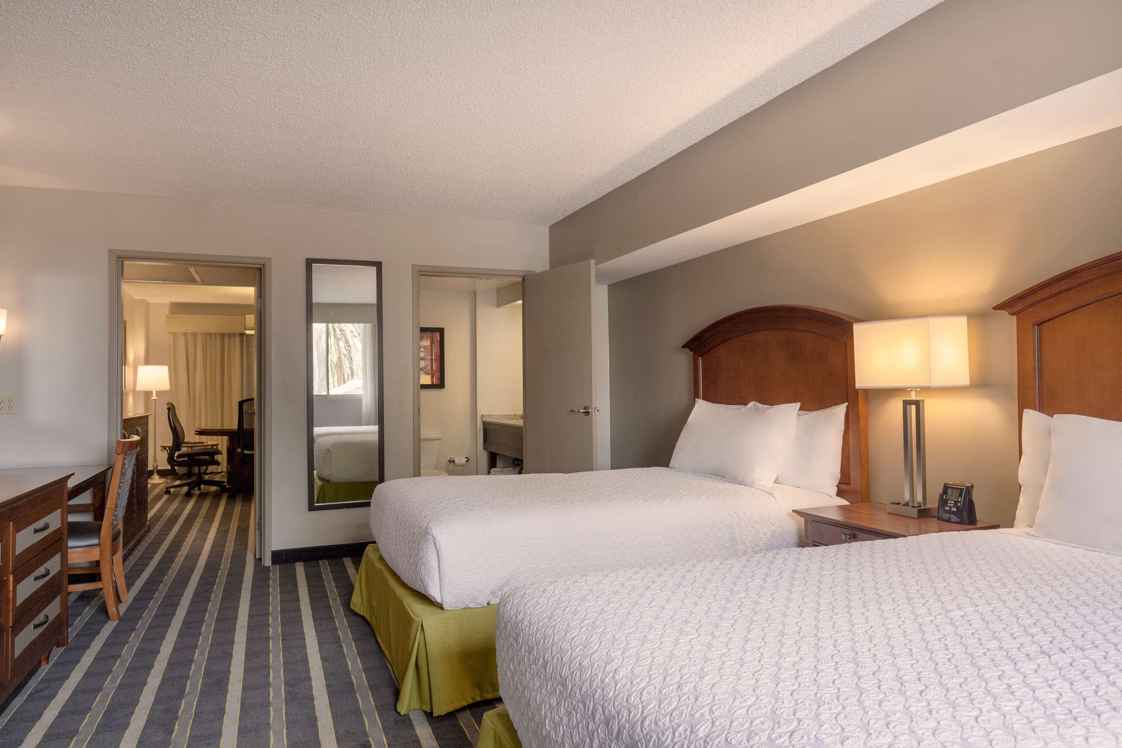 A hotel room with two beds, a desk, a chair, a mirror, and a nightstand with a lamp.