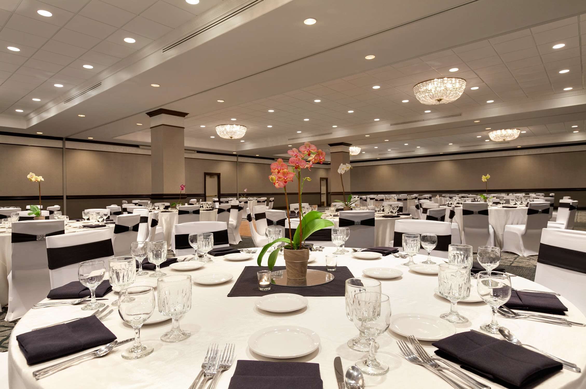 Un salón de banquetes con mesas redondas dispuestas para el servicio de comidas, cada una con mantel blanco, fundas negras para las sillas y cubertería de vidrio.