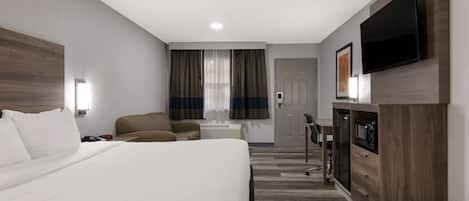 A hotel room with a large bed, a TV mounted on the wall, a desk with a chair, and a kitchenette area.