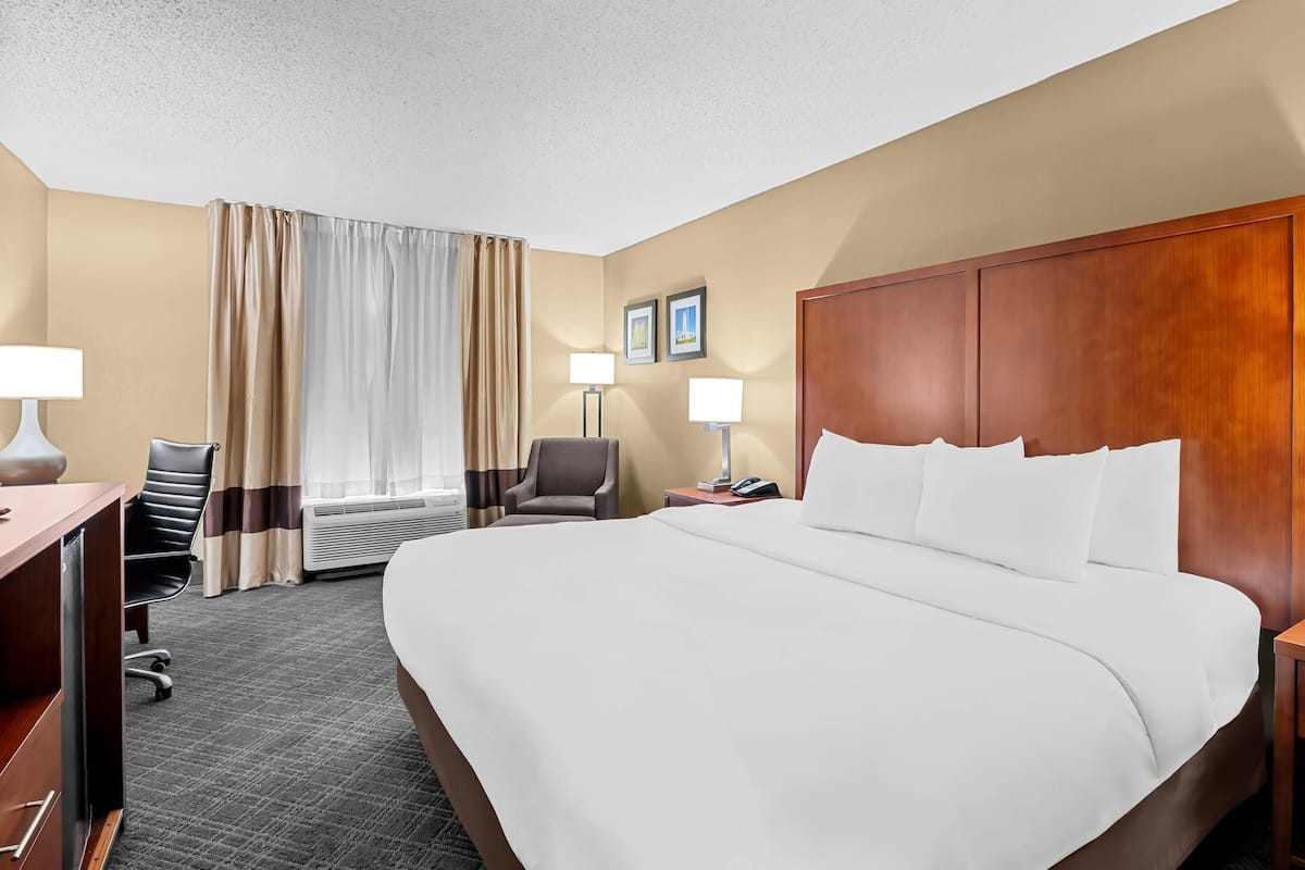 A hotel room with a large bed, a desk with a chair, a desk lamp, a bedside table with a lamp, and a window with curtains.