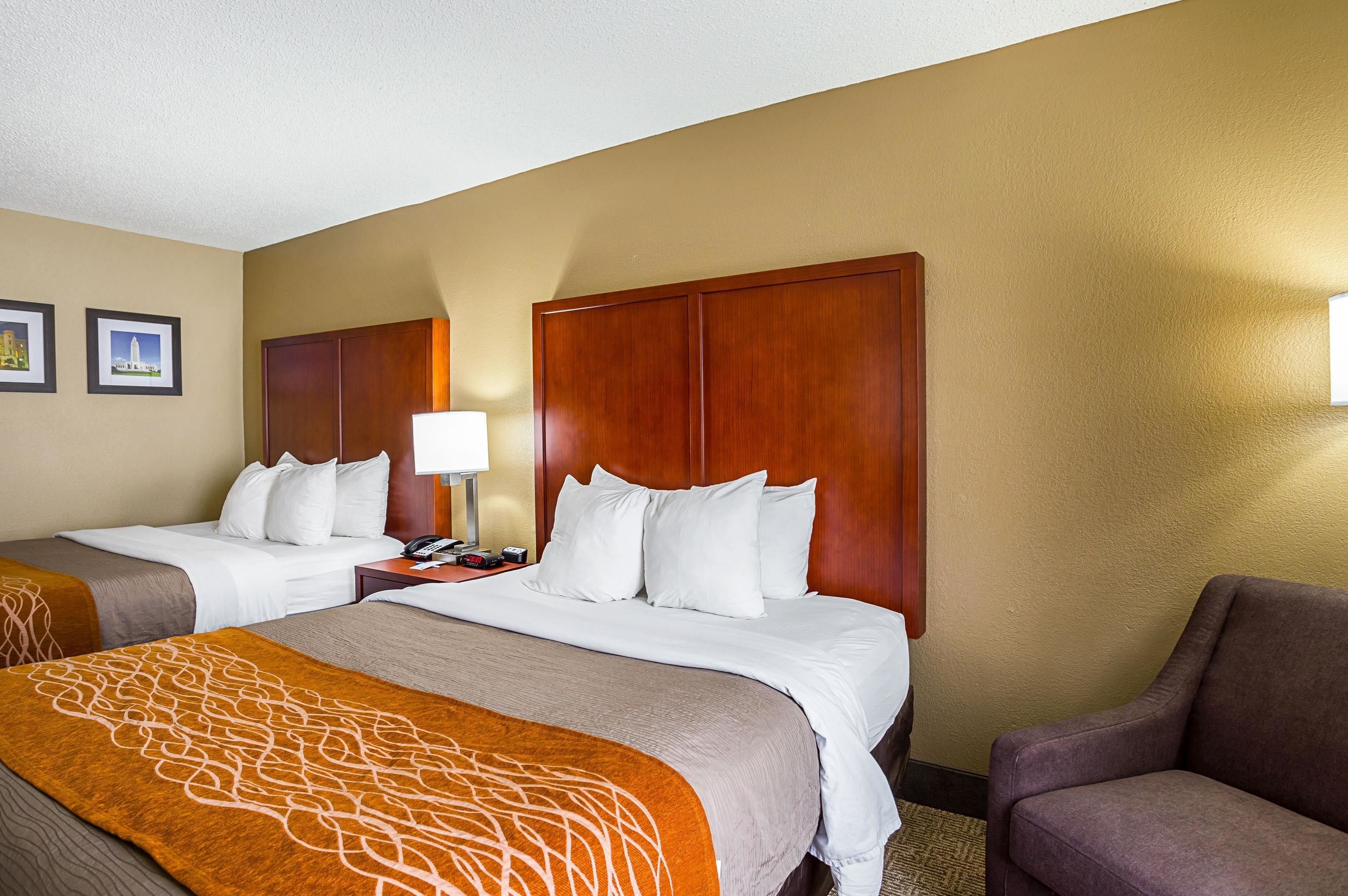 A hotel room with two beds, a wooden headboard, a nightstand with a lamp, and a brown armchair.