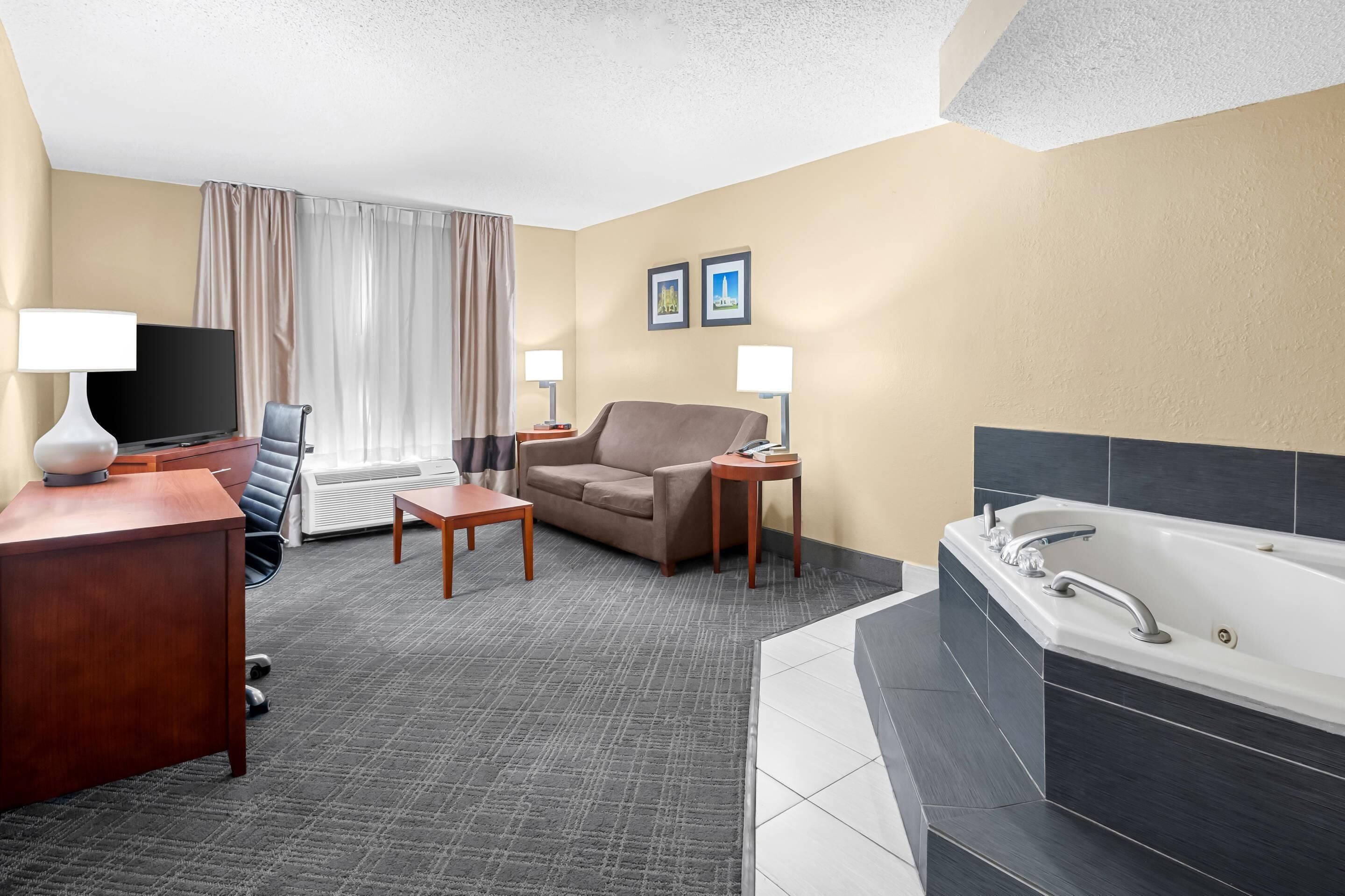 A hotel room with a sofa, a TV, a desk, a lamp, a small table, a bathroom with a bathtub, and two framed pictures on the wall.