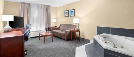 A hotel room with a sofa, a TV, a desk, a lamp, a small table, a bathroom with a bathtub, and two framed pictures on the wall.