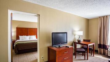 A hotel room with a bed, a TV on a wooden dresser, a desk with a lamp, and a view of the hallway.