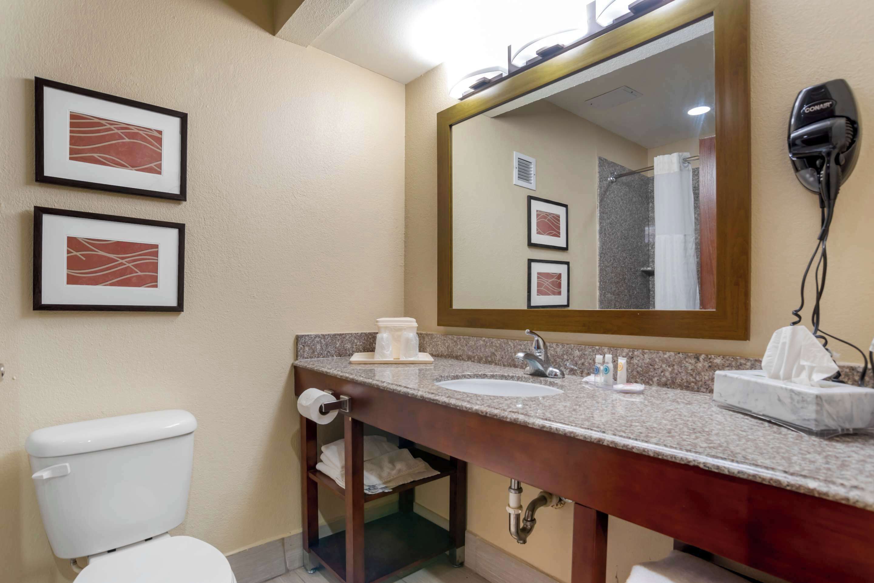 A bathroom with a toilet, sink, and large mirror. There are framed pictures on the wall and a hair dryer on the wall.