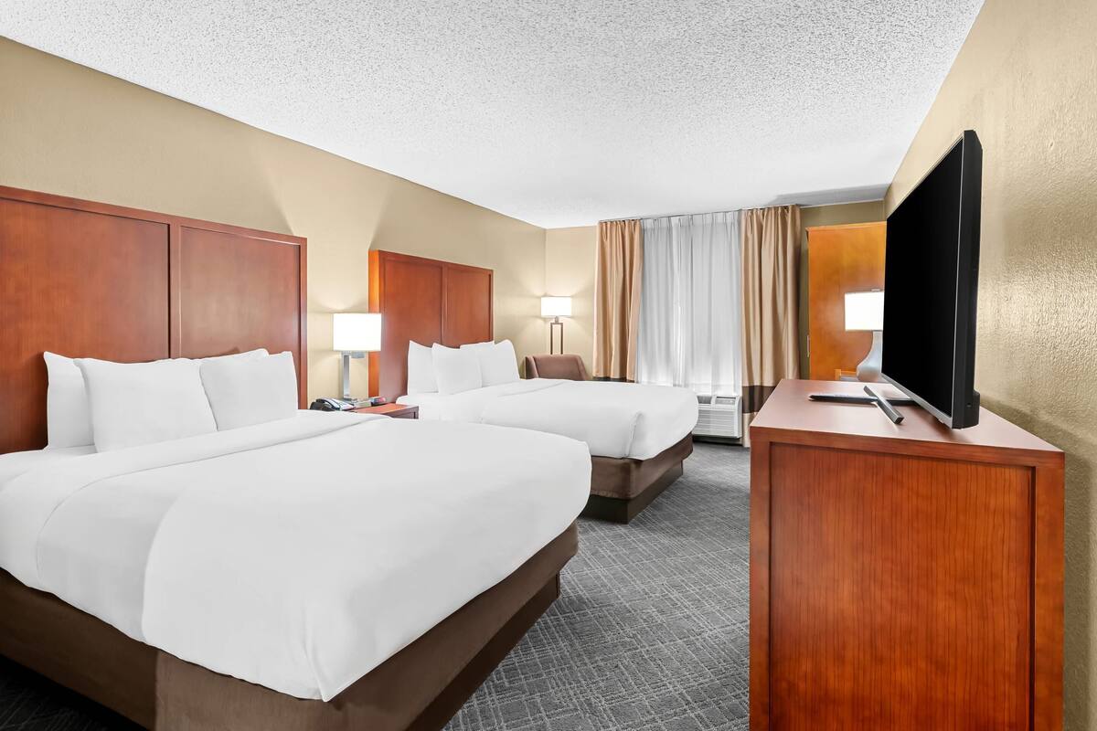 A hotel room with two beds, a desk, a TV, and a large window with curtains.