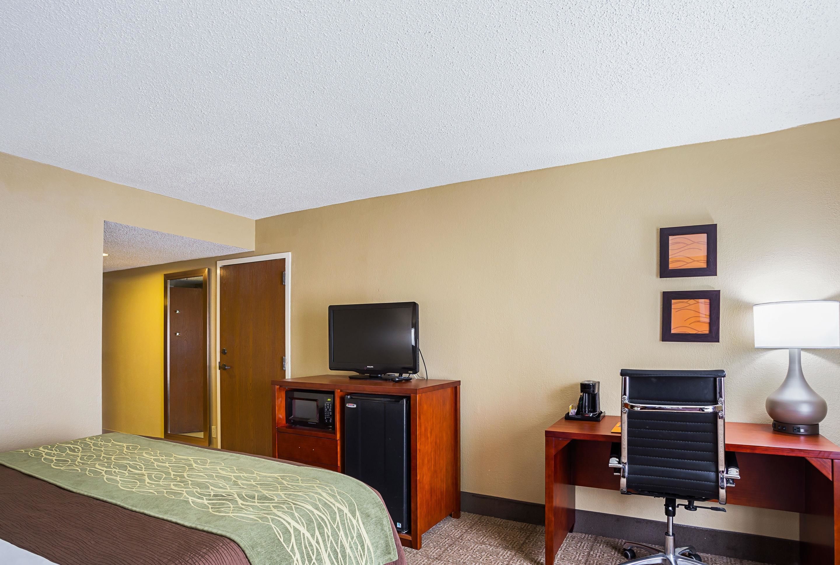 Een hotelkamer met een bed, een bureau met een televisie, een kleine koelkast, een bureaulamp en een fotolijst aan de muur.