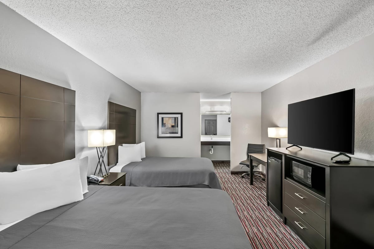 A hotel room with two beds, a desk, a TV, a microwave, and a bathroom visible through an open door.