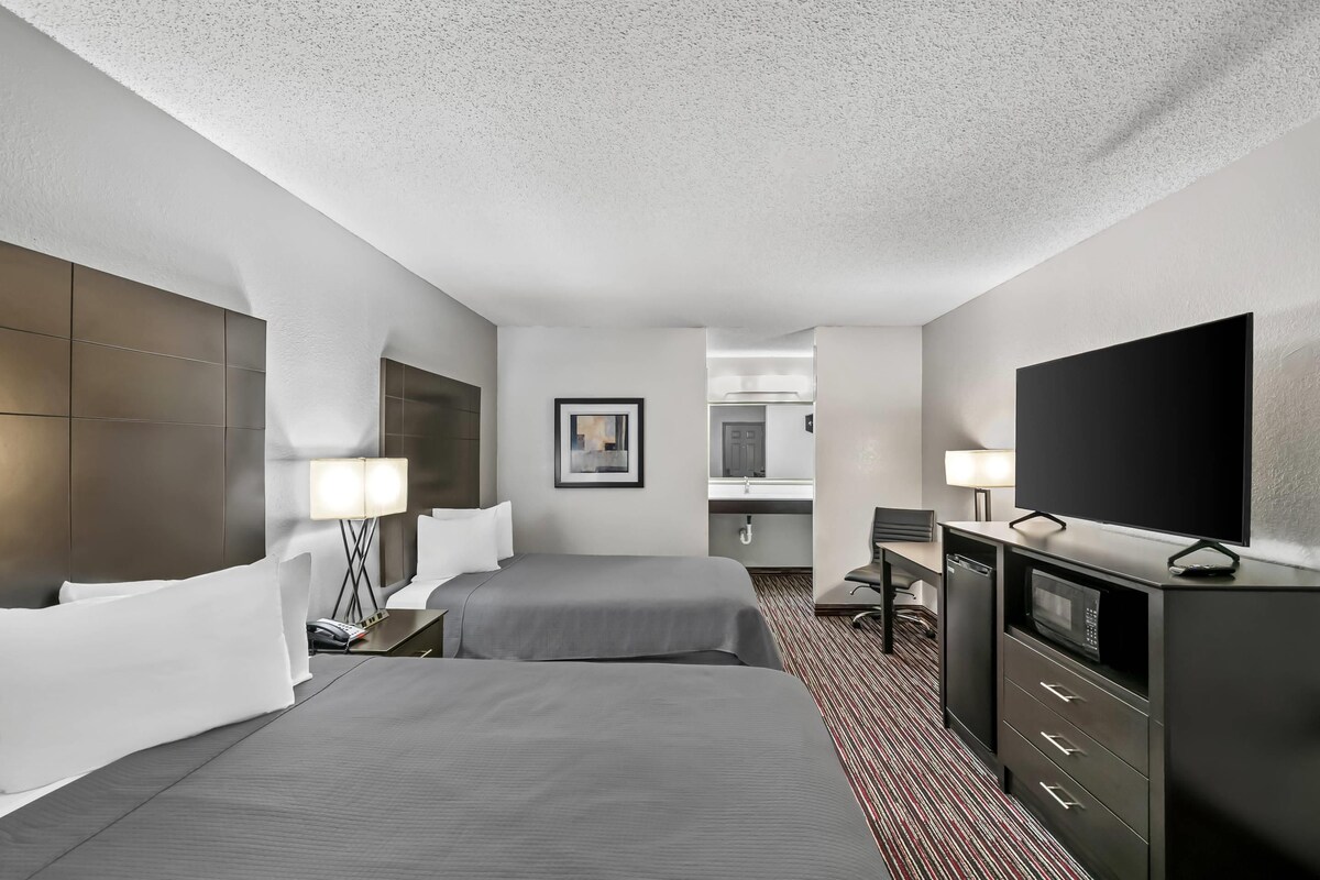 A hotel room with two beds, a desk, a TV, a microwave, and a bathroom visible through an open door.