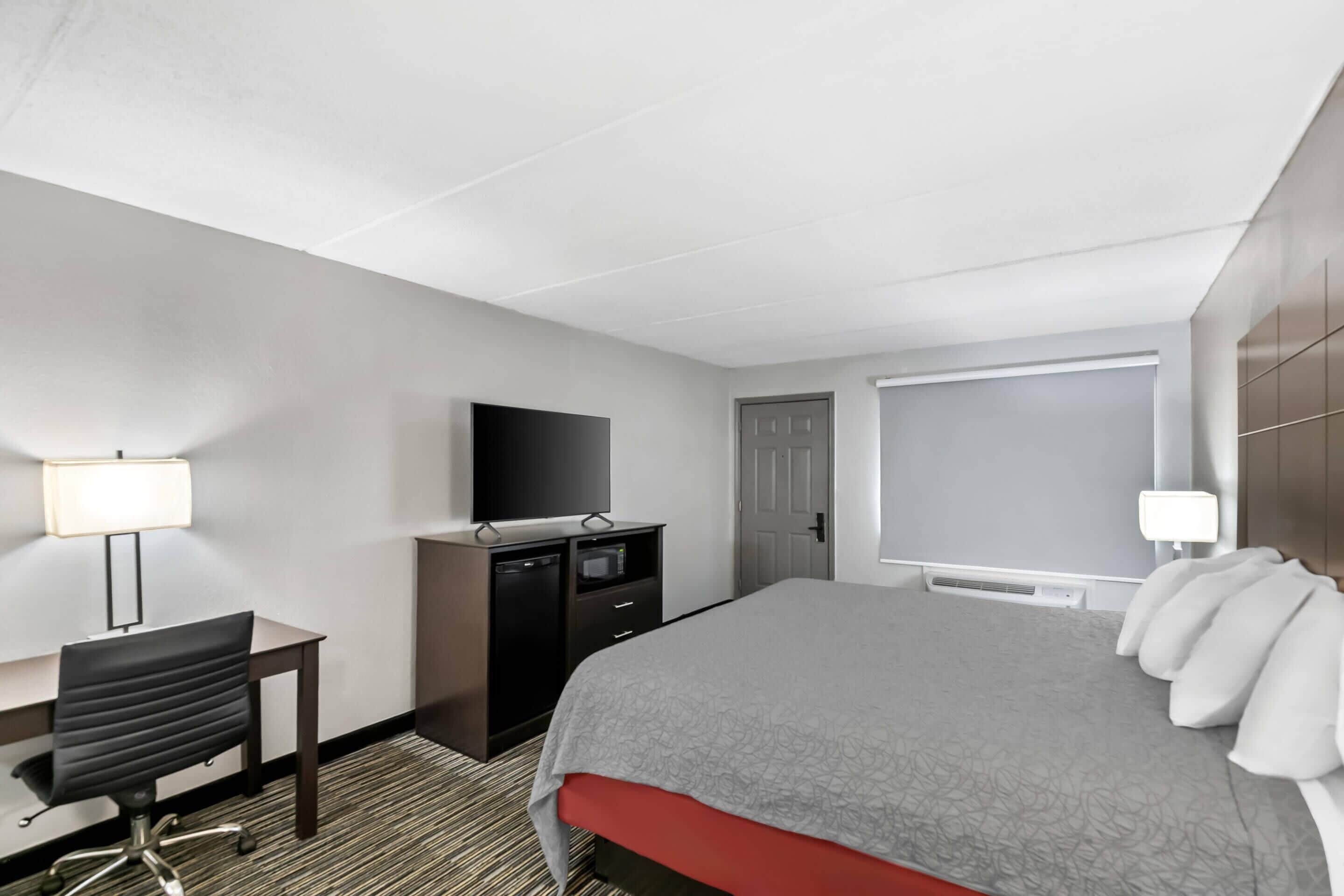 A hotel room with a bed, a desk with a chair, a television, a wardrobe, and a window with blinds.