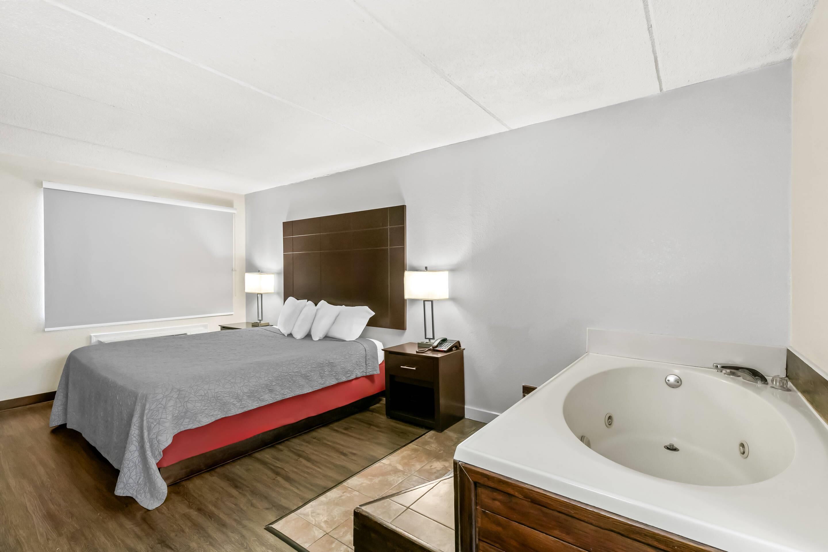 A hotel room with a bed, bedside table, lamp, and a large bathtub.