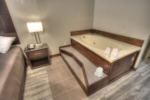 Habitación de hotel con una bañera grande, una mesita de noche con lámpara y una cama con almohada.