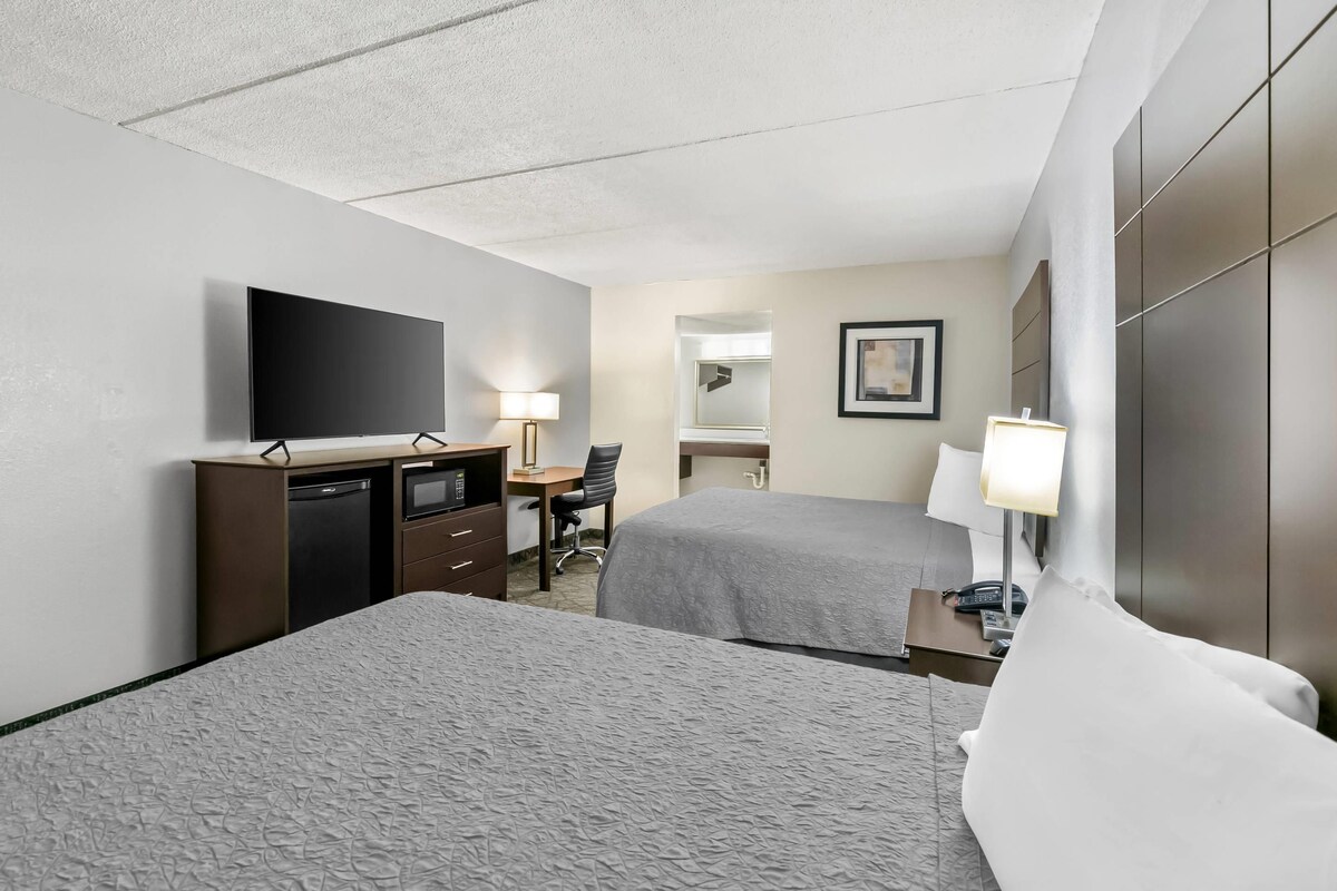 A hotel room with two beds, a desk with a chair, a flat-screen TV, a desk lamp, a mirror, and a bedside table with a lamp.