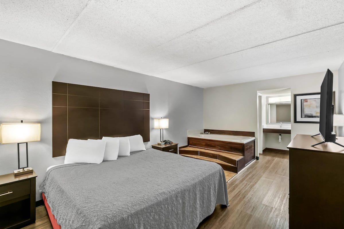 A hotel room with a large bed, a wooden headboard, a desk with a lamp, a bathroom with a sink and mirror, and a TV on a stand.