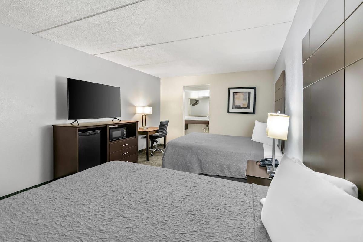 A hotel room with two beds, a desk with a chair, a flat-screen TV, a desk lamp, and a bedside table with a lamp.