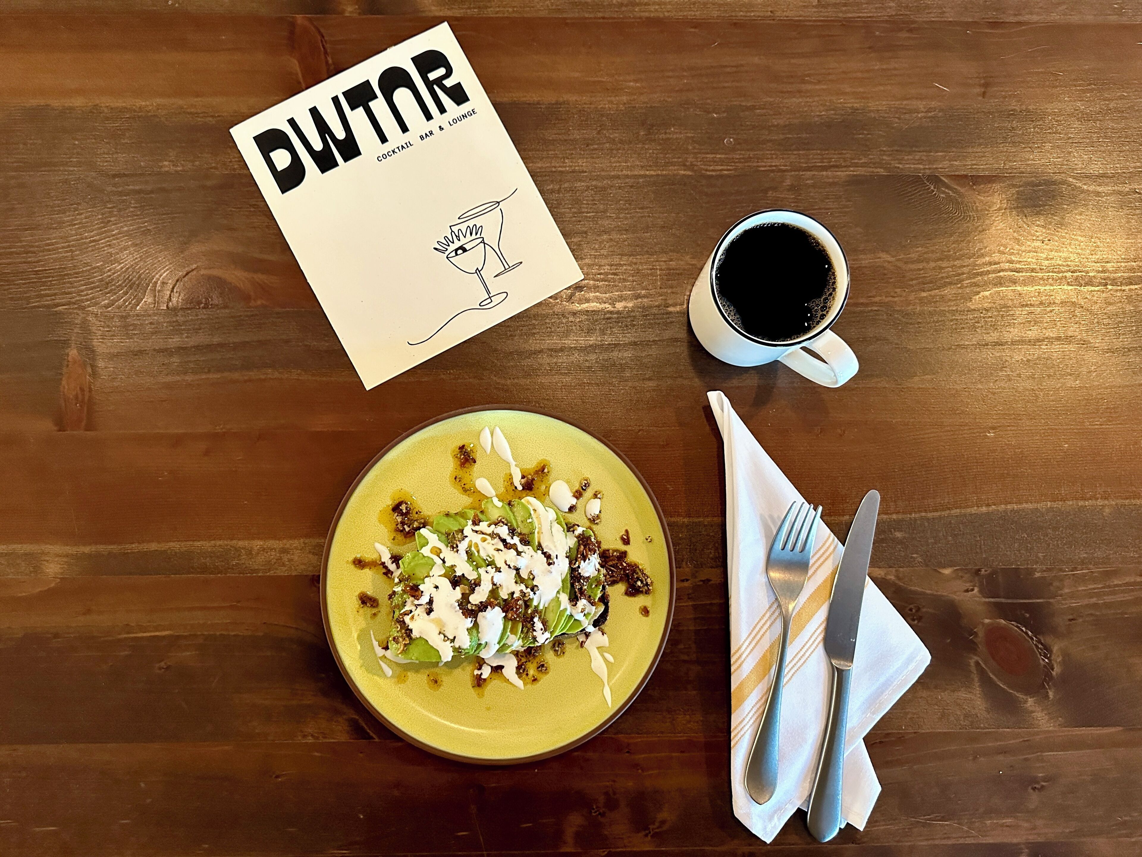 A wooden table with a yellow plate of food, a cup of black coffee, and a menu titled "DWNTAR".