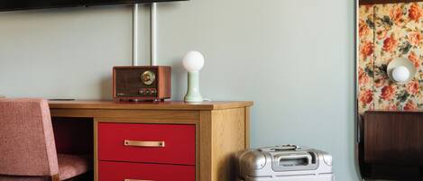 A room with a wooden desk, a red drawer unit, a vintage radio, a small lamp, a silver suitcase, a television mounted on the wall, and a mirror with a floral wallpaper pattern.
