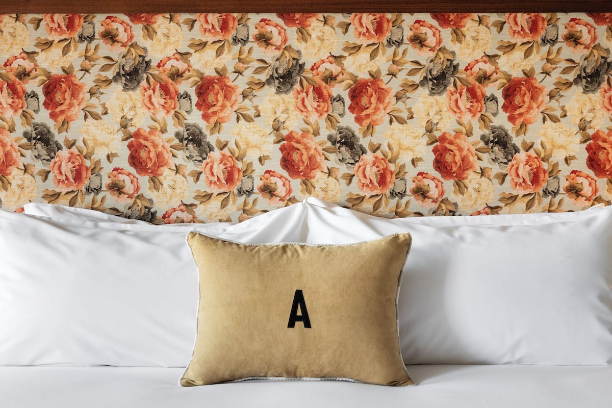 A bed with white linens and a beige pillow with a large 'A' on it, set against a floral wallpaper backdrop.