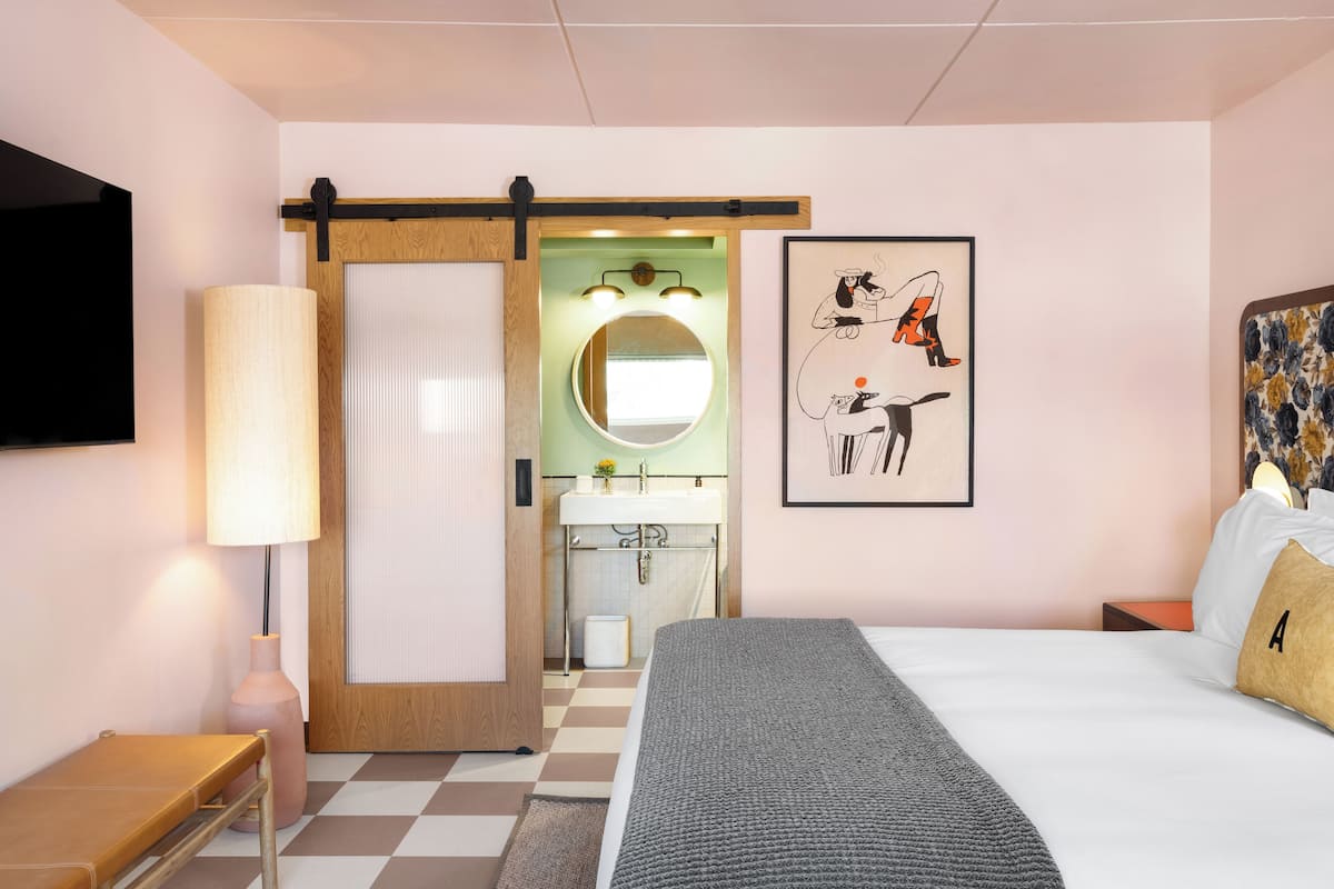 A hotel room with a bed, a bench, a lamp, a painting, and a bathroom visible through an open door.