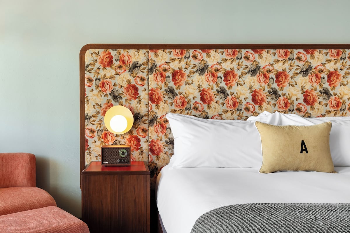 A hotel room with a floral-patterned headboard, a bedside table with a radio, a lamp, a bed with white bedding and a pillow with the letter 'A', and a grey textured rug.