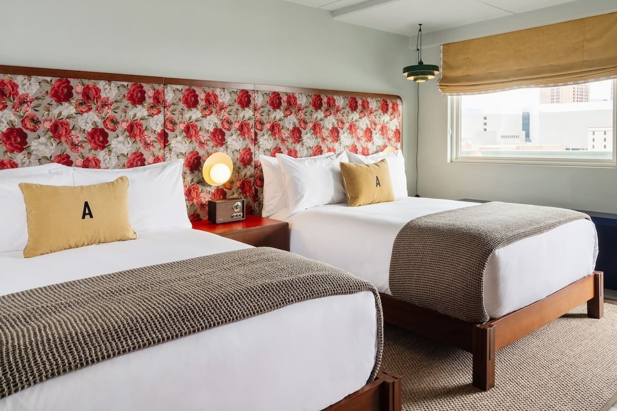 A hotel room with two beds, floral wallpaper, a nightstand with a lamp, and a window with a view of buildings.