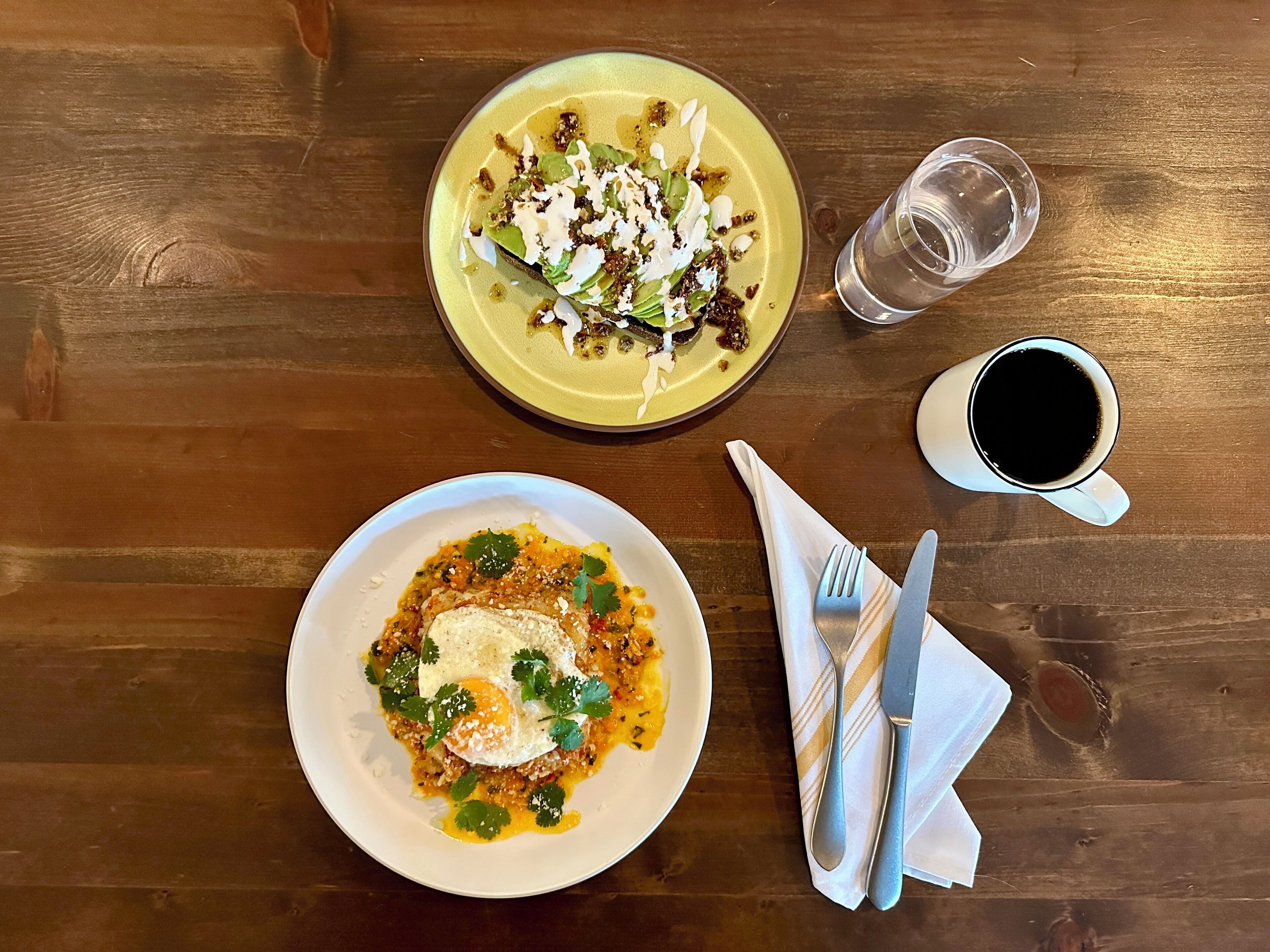 Two plates of food on a wooden table with a glass of water and a cup of coffee.