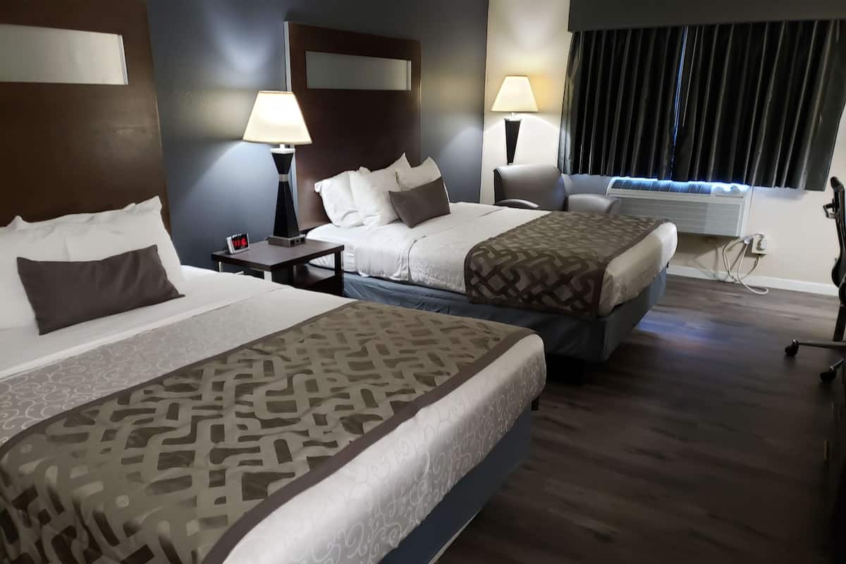 A hotel room with two beds, a nightstand with a lamp, a window with curtains, and a wall-mounted air conditioner.