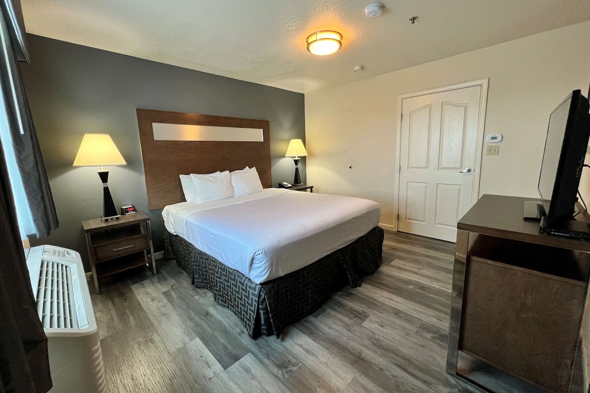 A hotel room with a large bed, two bedside tables with lamps, a desk with a computer, and a door.