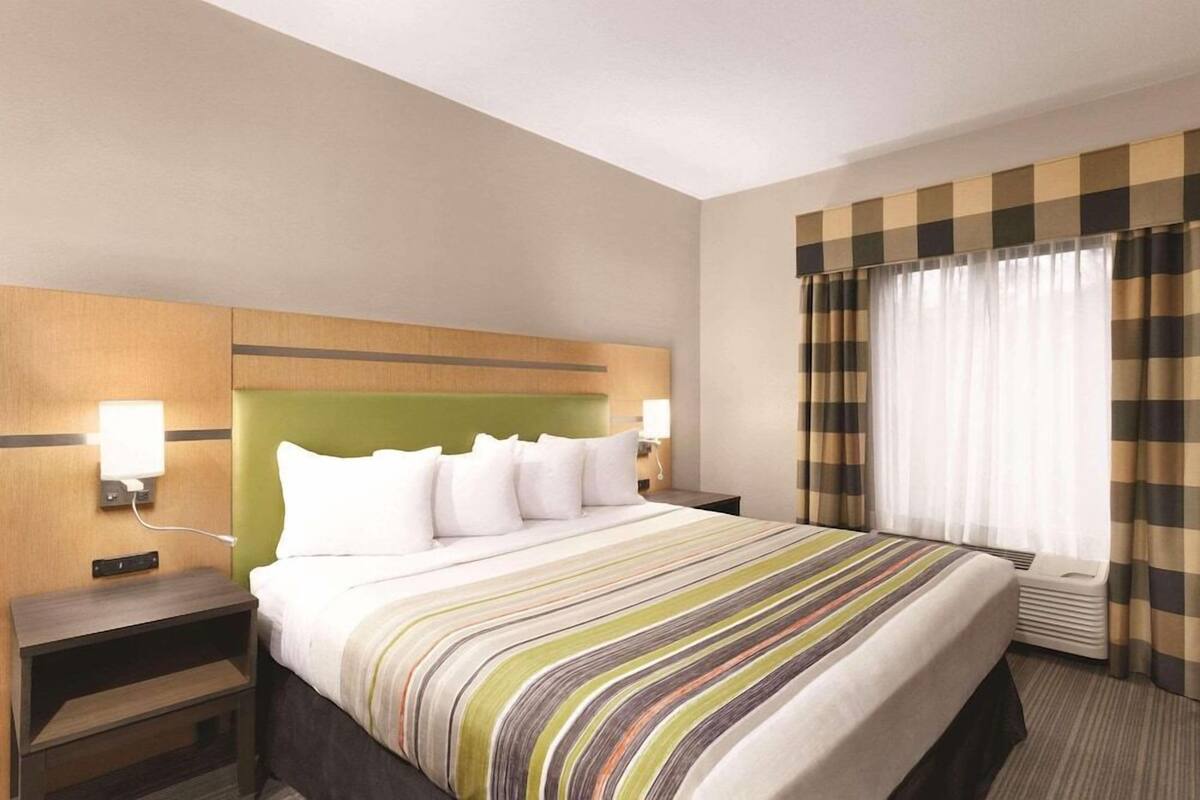 A hotel room with a bed, a nightstand, a lamp, a window with curtains, and a wall-mounted air conditioning unit.