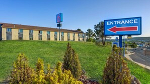 A hotel entrance sign with a directional arrow, a building with multiple windows, and a grassy area with shrubs. - Motel 6 Silver City, NM (Silver City)