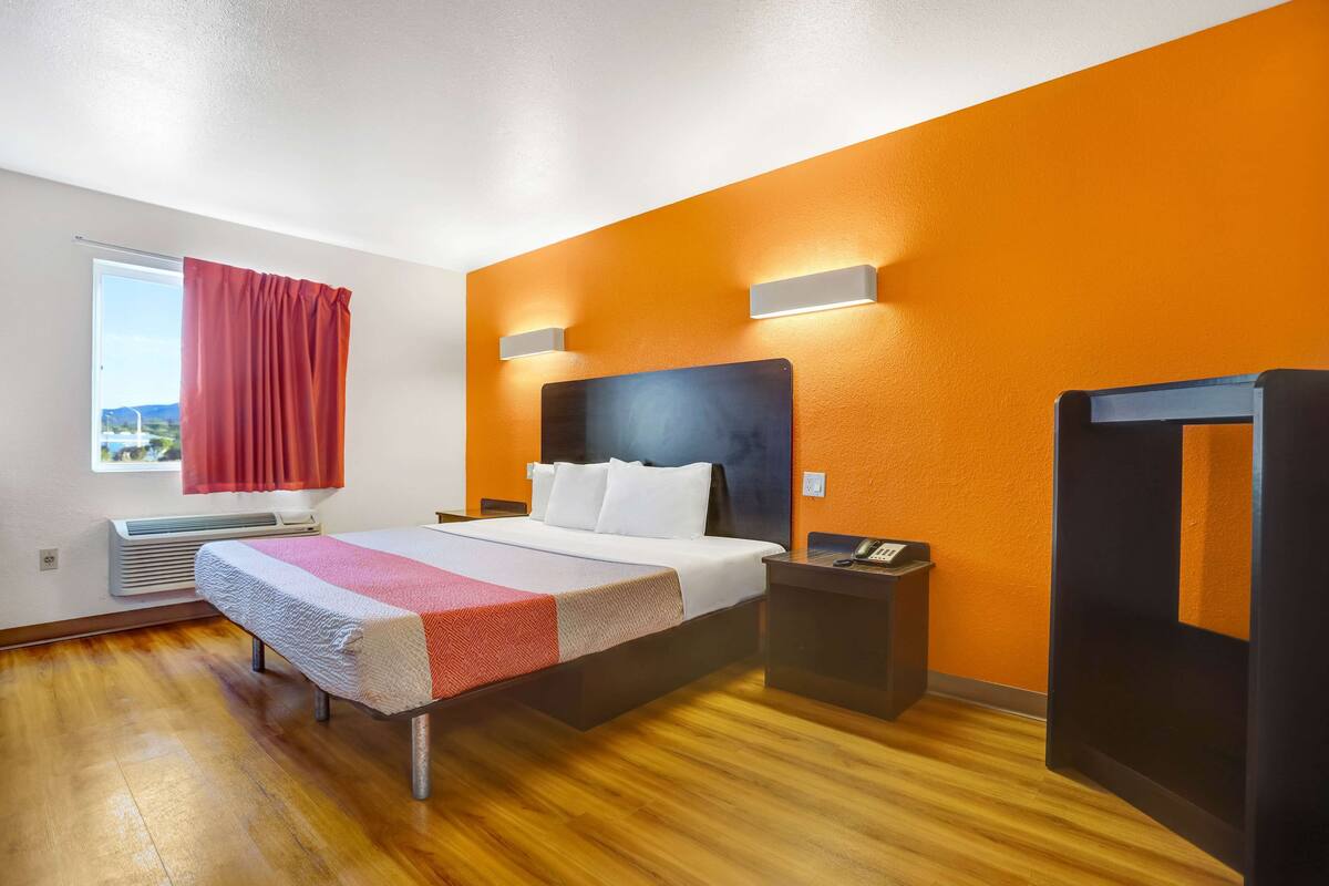 A hotel room with a bed, a nightstand, a desk, a window with curtains, and an air conditioning unit.