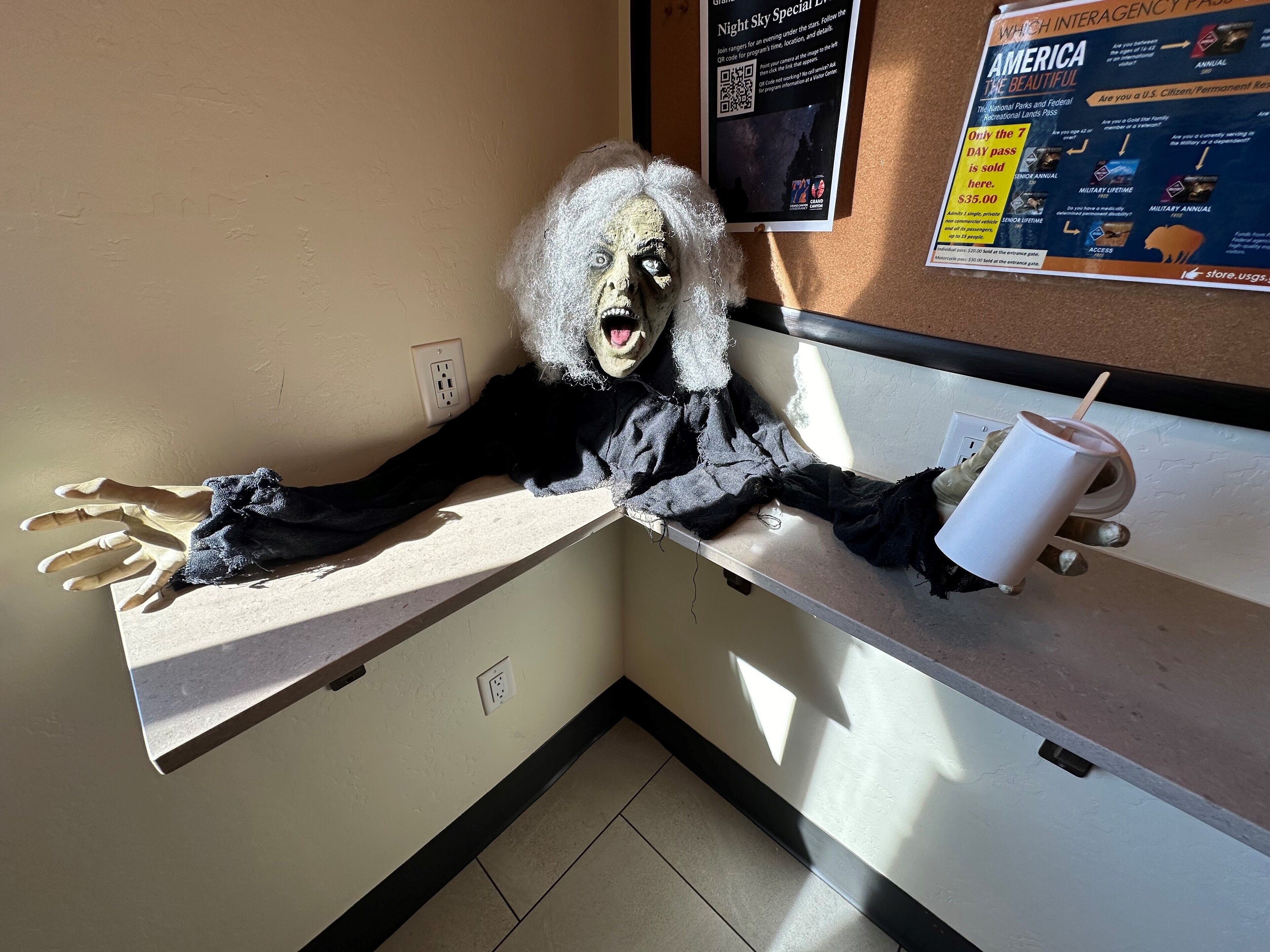 A mannequin with a white wig and black clothing, positioned on a counter with a white cup and straw.
