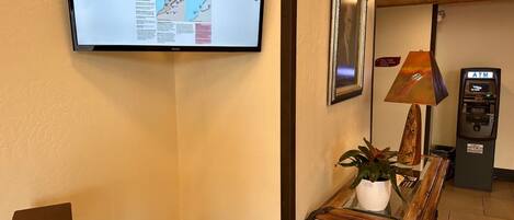 A hotel lobby with a digital display, a framed picture, an ATM, a lamp, a plant, and an express check-out box.