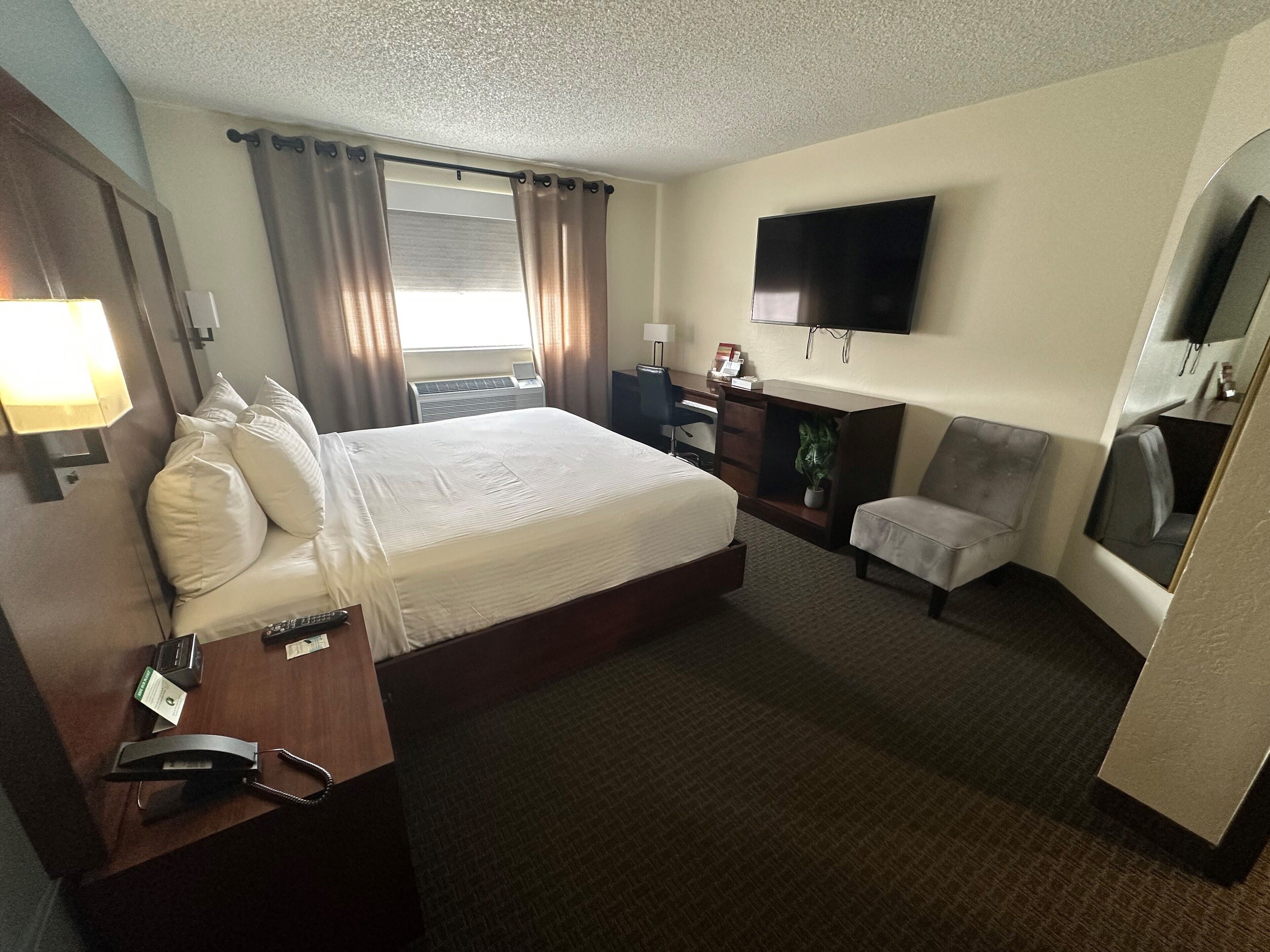 A hotel room with a large bed, a desk with a chair, a TV mounted on the wall, and a window with curtains.