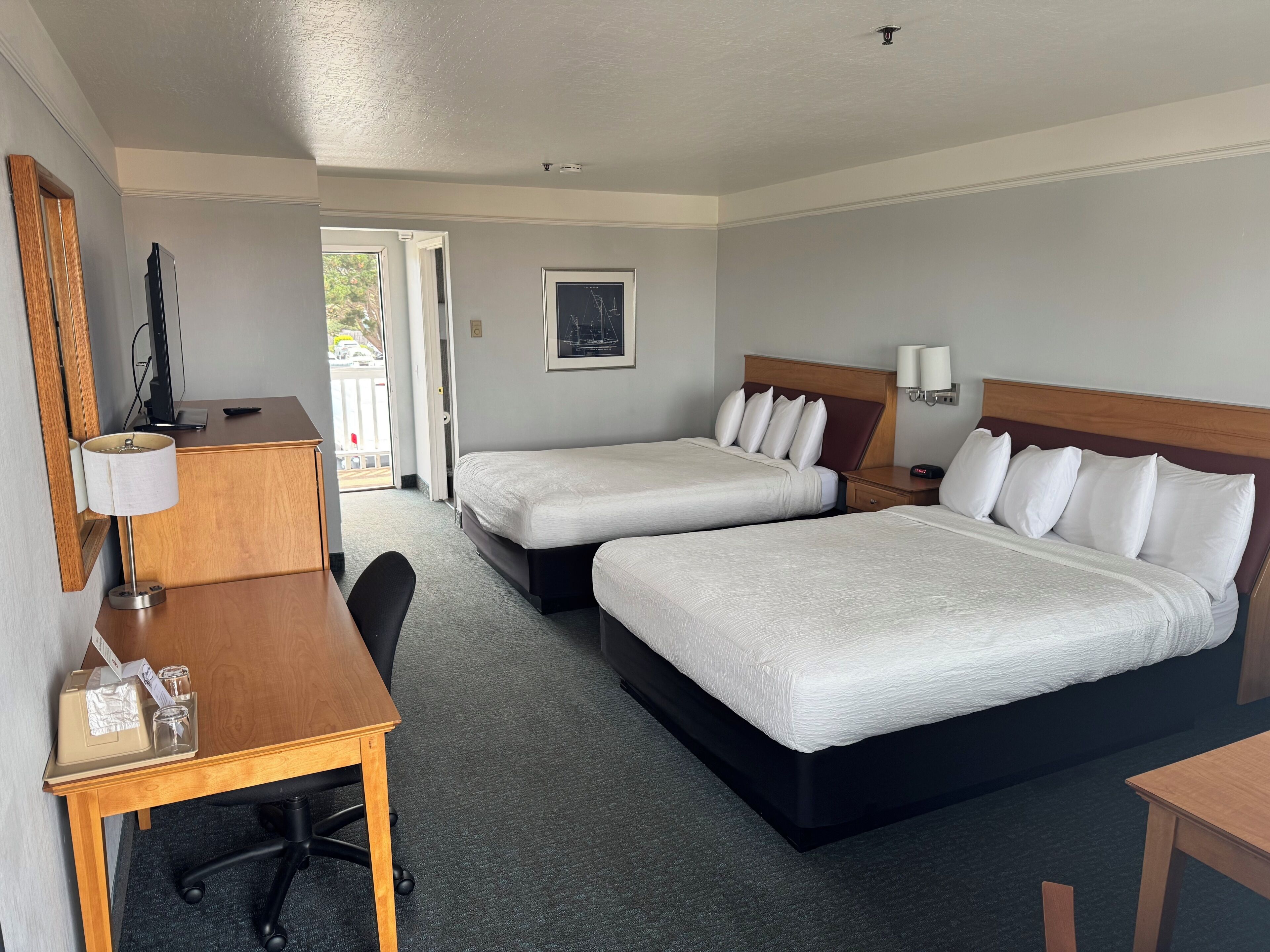 A hotel room with two beds, a desk with a chair, a television, and a door leading to an outdoor area.