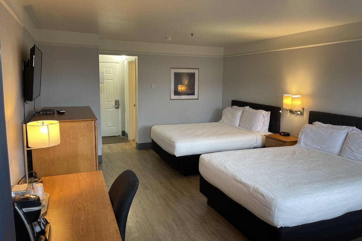 A hotel room with two beds, a desk with a lamp, a coffee machine, a TV, and a door leading to another room.