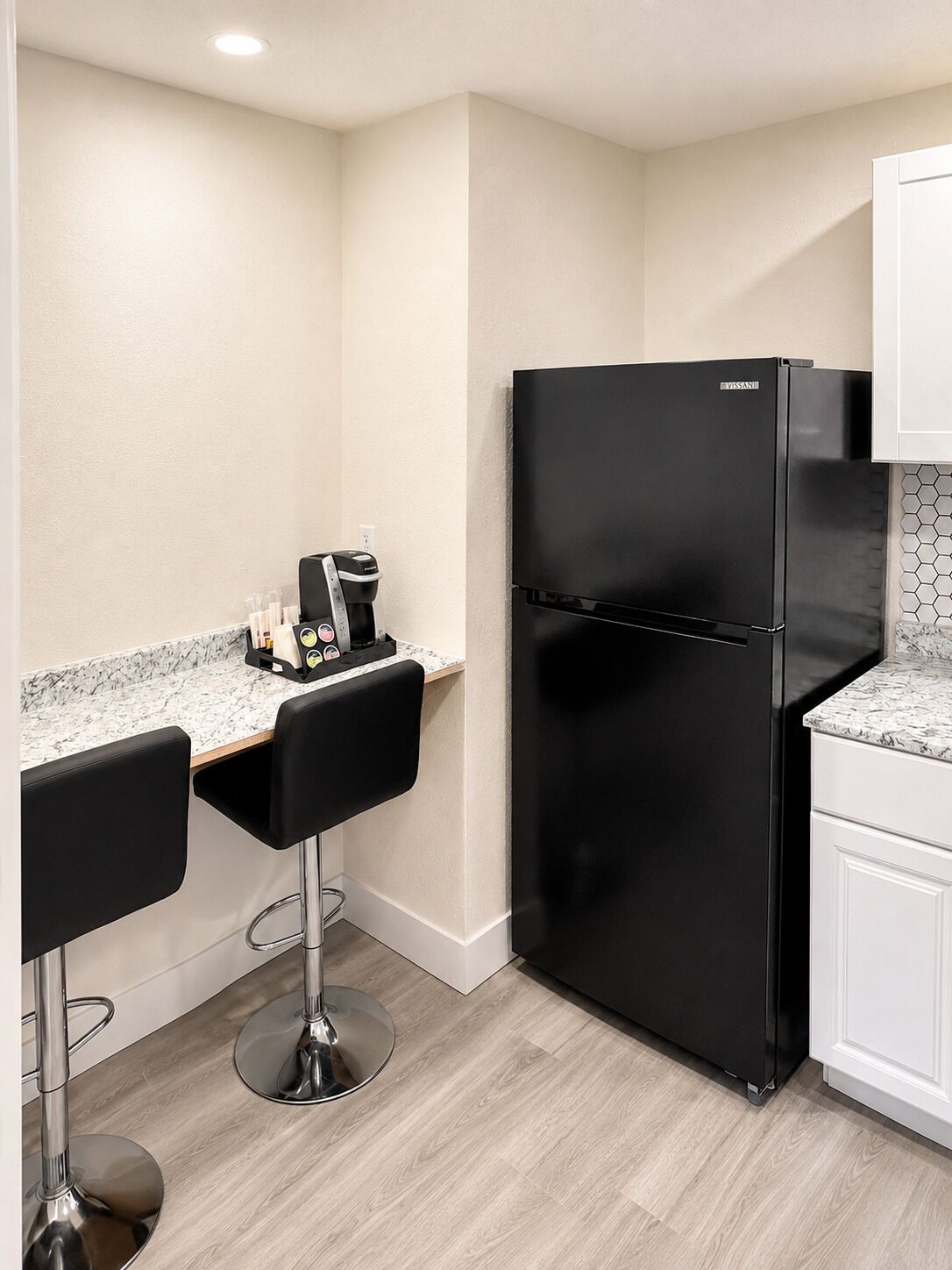 A compact kitchen with a black refrigerator, two bar stools, a coffee machine, and a marble countertop.