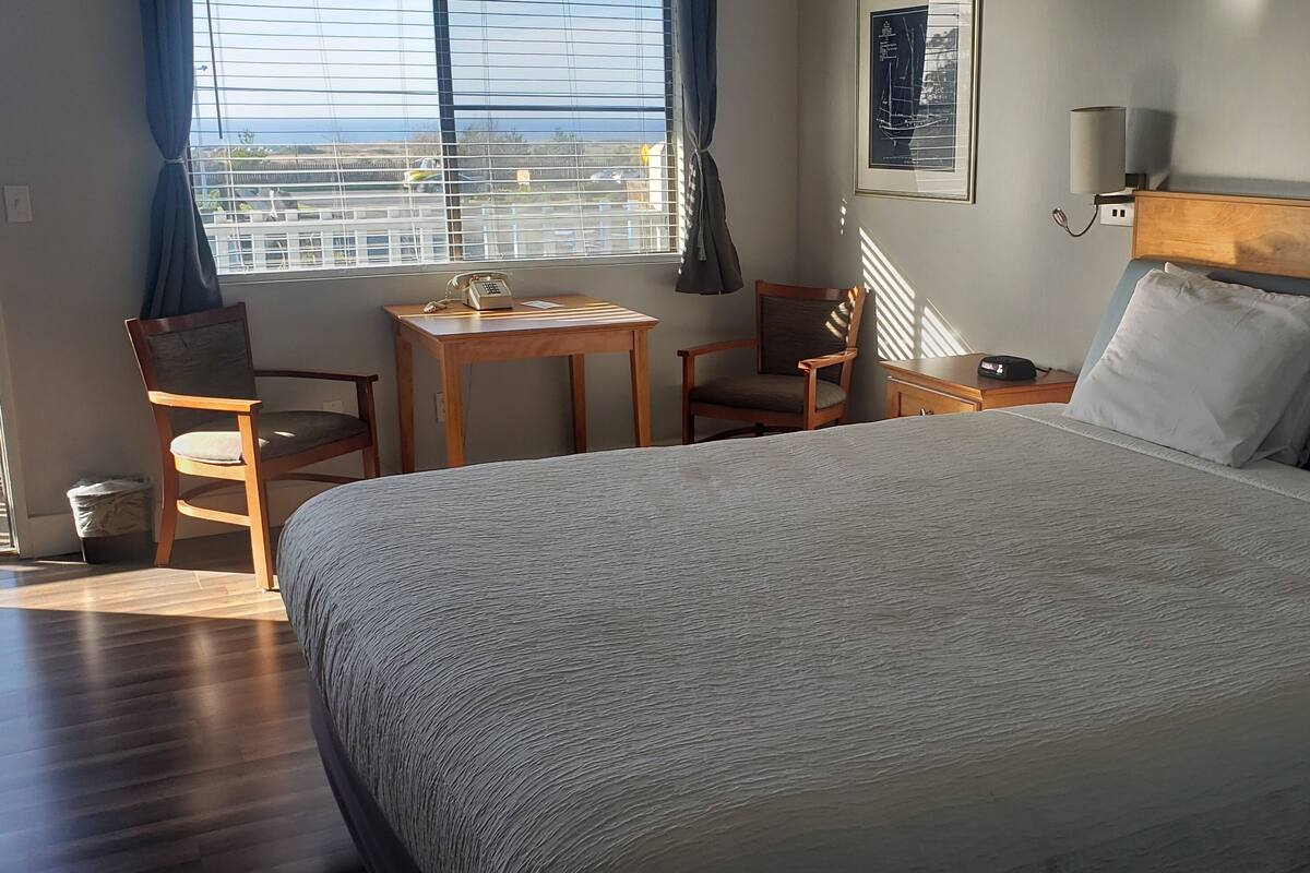A hotel room with a large bed, a desk, a chair, a small table, and a window with a view.