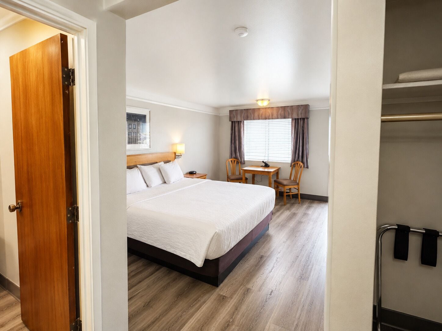 Een hotelkamer met een bed, een bureau, twee stoelen, een raam met gordijnen en een kapstok.
