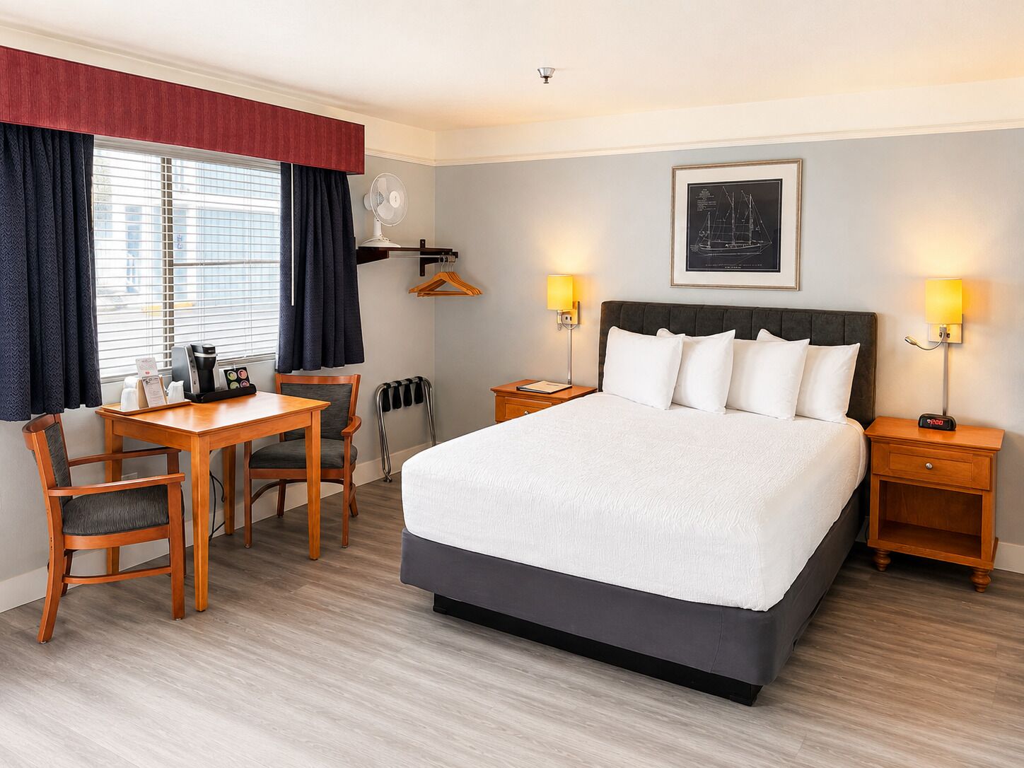 Habitación de hotel con cama, mesitas de noche, un escritorio con sillas, una ventana con cortinas y un cuadro colgado en la pared.