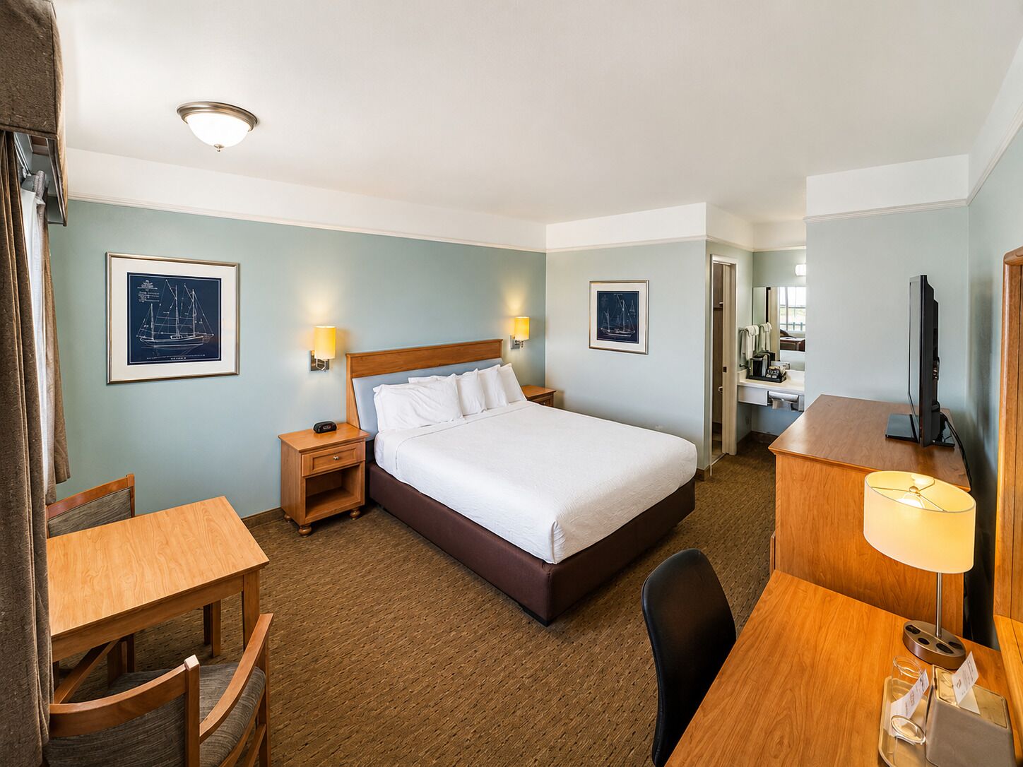 A hotel room with a bed, bedside tables, a desk, a chair, a television, and framed pictures on the walls.