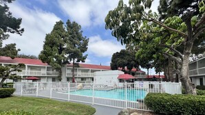 A pool area with a fenced deck, surrounded by a white fence, and a building with a red roof in the background. - Motel 6 Irvine - Orange County Airport (Santa Ana)