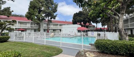 A pool area with a fenced deck, surrounded by a white fence, and a building with a red roof in the background.