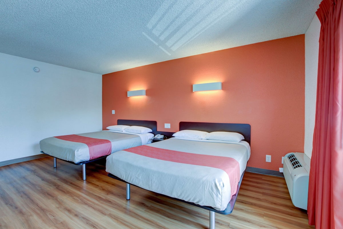 A hotel room with two beds, wooden flooring, a red wall, and a ceiling light fixture.