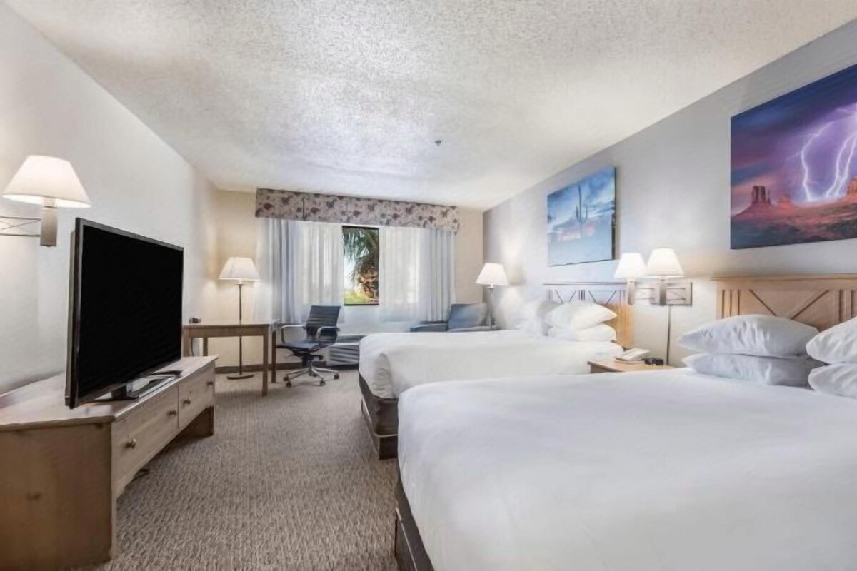 A hotel room with two beds, a TV, a desk, and a large window.